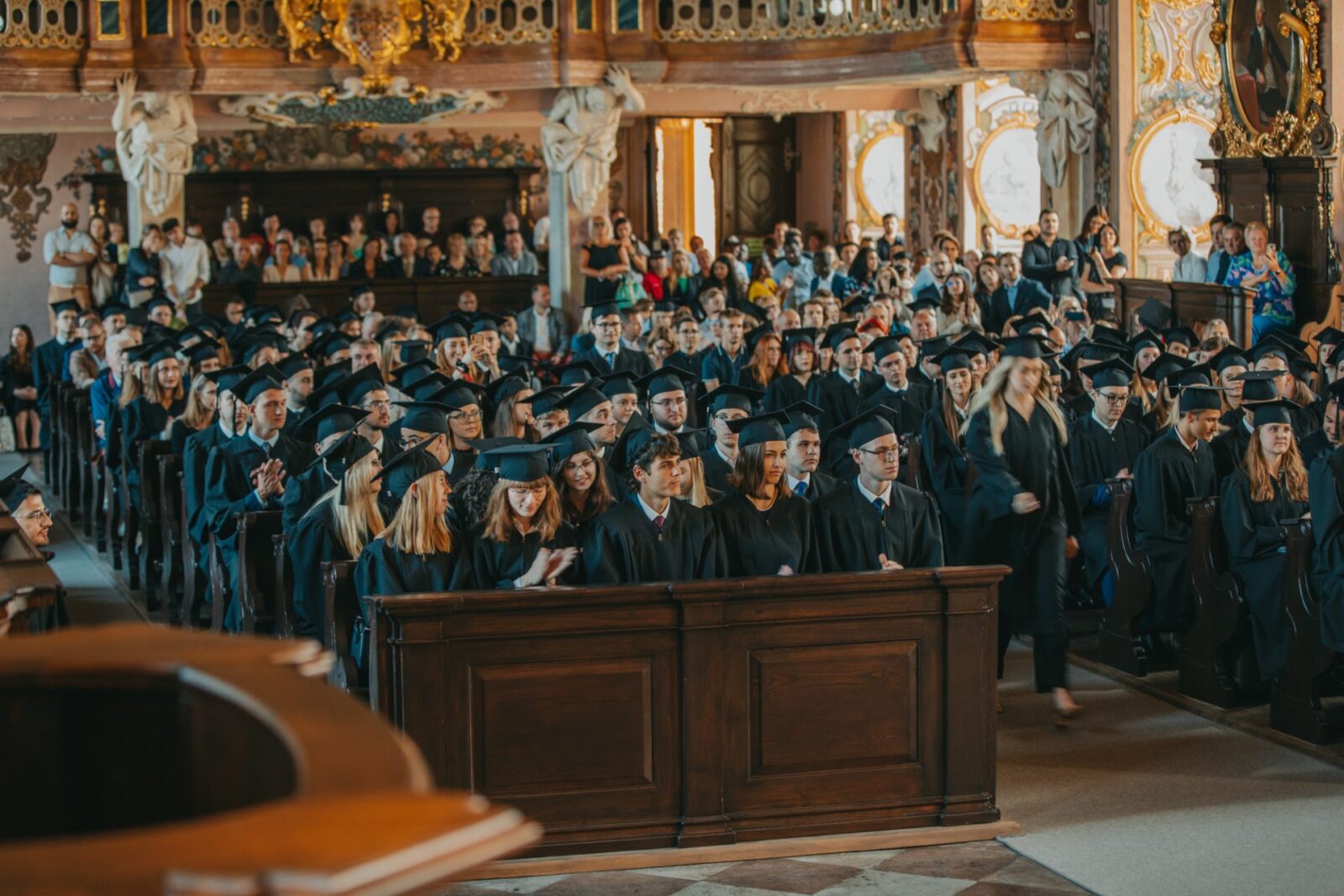 absolwenci w togach i biertach w zabytkowej sali